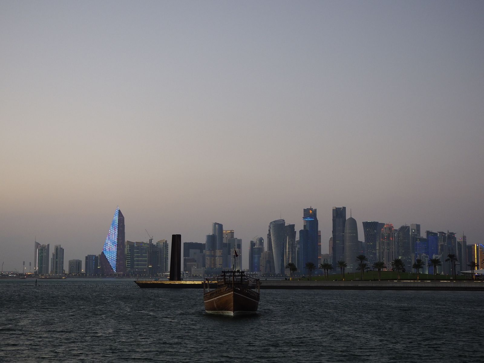 Scenic view of Doha city skyline with traditional dhow boat at dusk along the waterfront.
