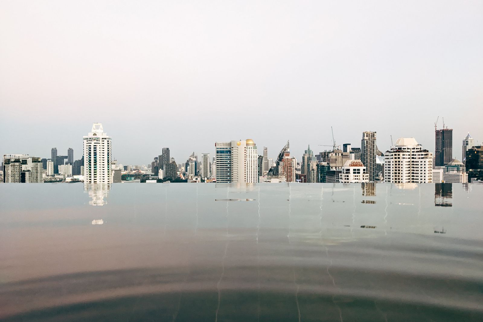 Tranquil infinity pool with reflections of the Bangkok skyline under a calm sky.